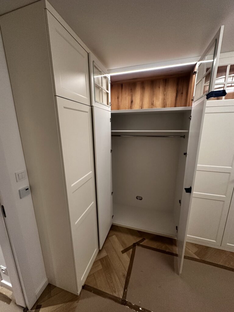 Biała szafa wnękowa z półkami i drewnianym panelem u góry.
