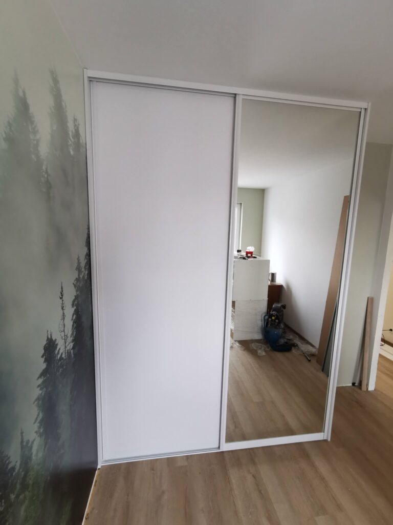 Biała szafa przesuwna z dużym lustrem przy leśnej fototapecie