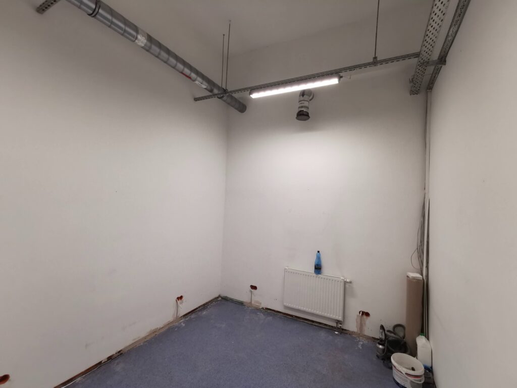 Pusty biały pokój z niebieską podłogą i widocznymi przewodami pod sufitem.