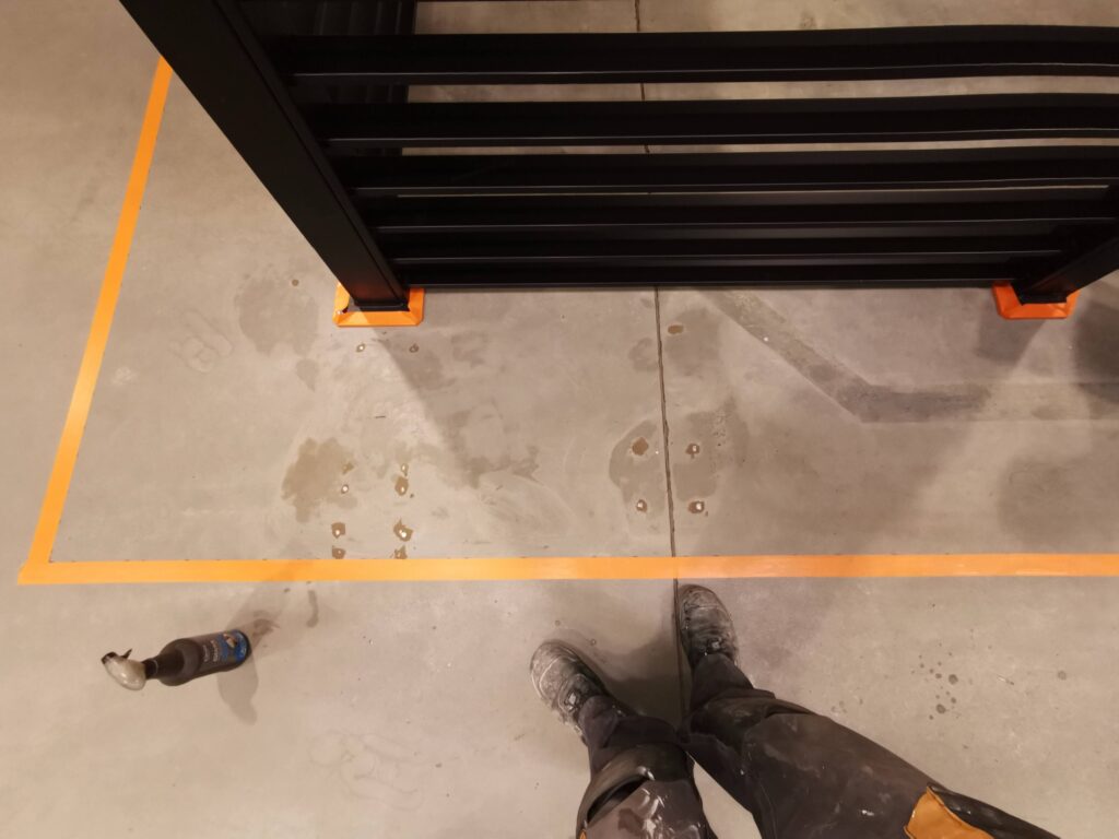 Czarna metalowa przegroda na betonowej podłodze, obok widoczne buty.