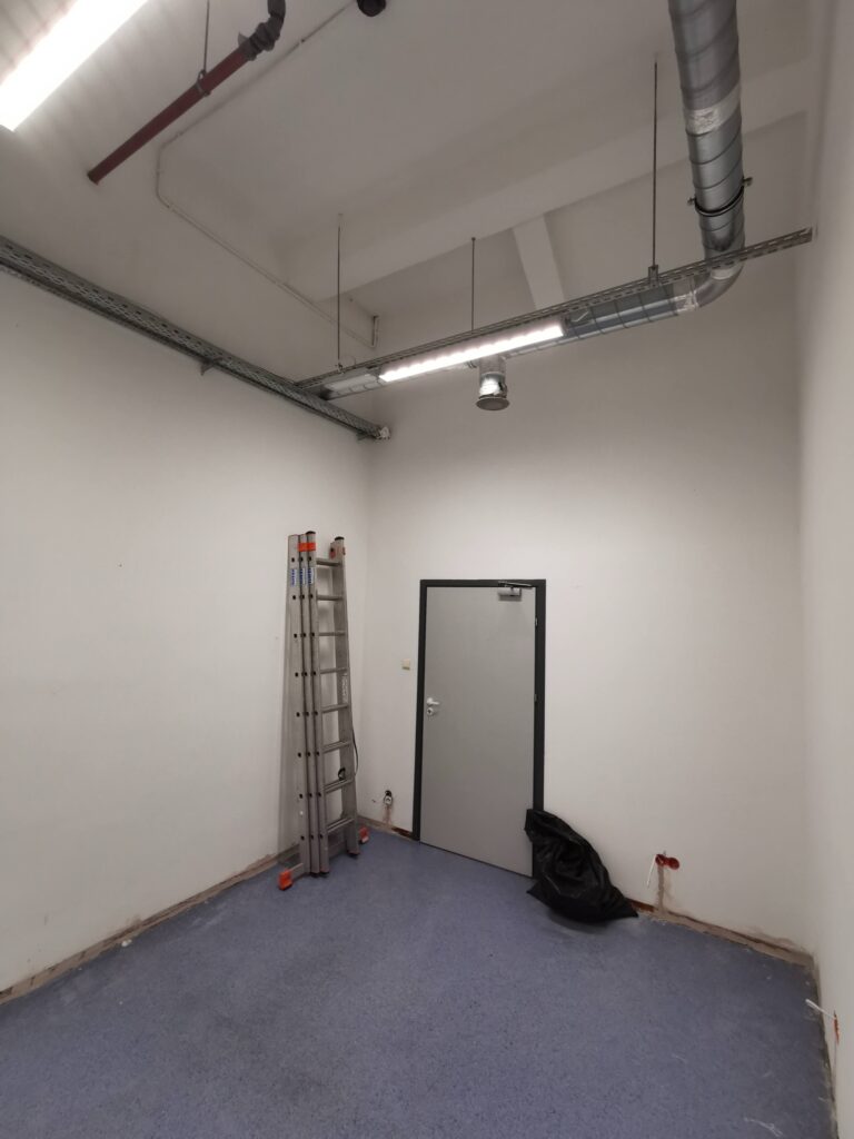 Puste pomieszczenie z niebieską podłogą, drabiną i instalacją pod sufitem.