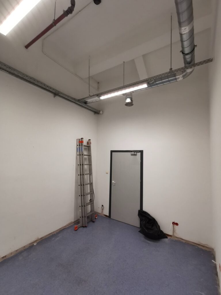 Puste białe pomieszczenie z drabiną i instalacjami pod sufitem.