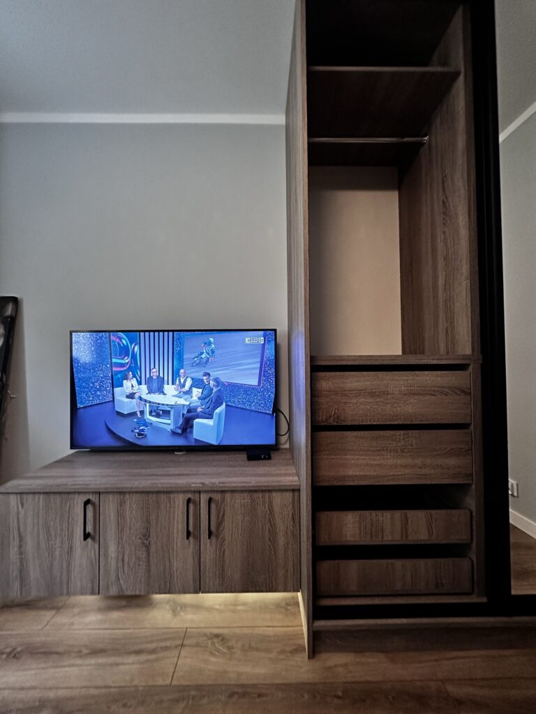 Telewizor nad drewnianą szafką i wysoka zabudowa z półkami.