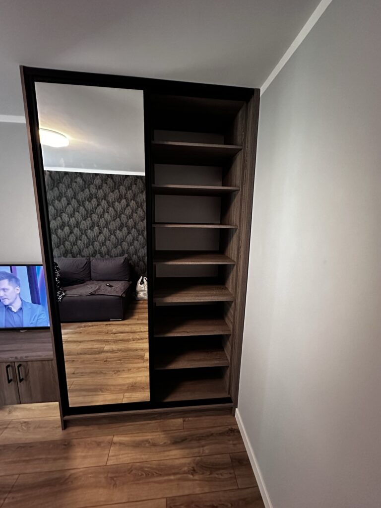 Ciemna zabudowa z półkami i lustrem obok telewizora.