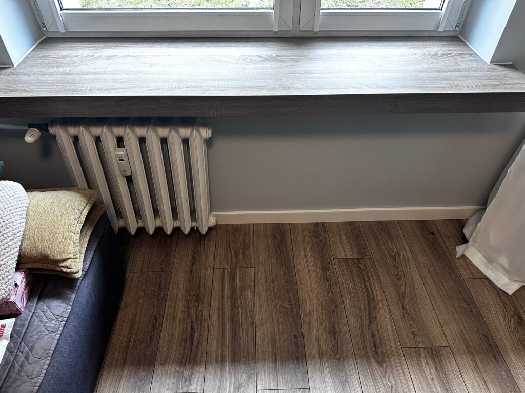 Grzejnik pod oknem nad drewnianą podłogą.