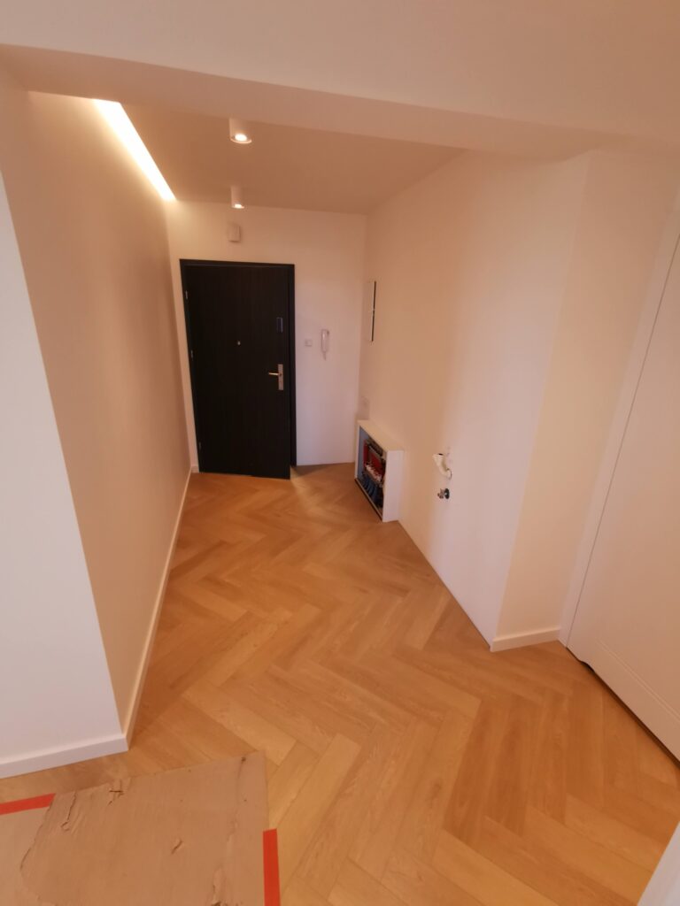 Jasny korytarz z parkietem, ciemnymi drzwiami i odsłoniętymi przyłączami