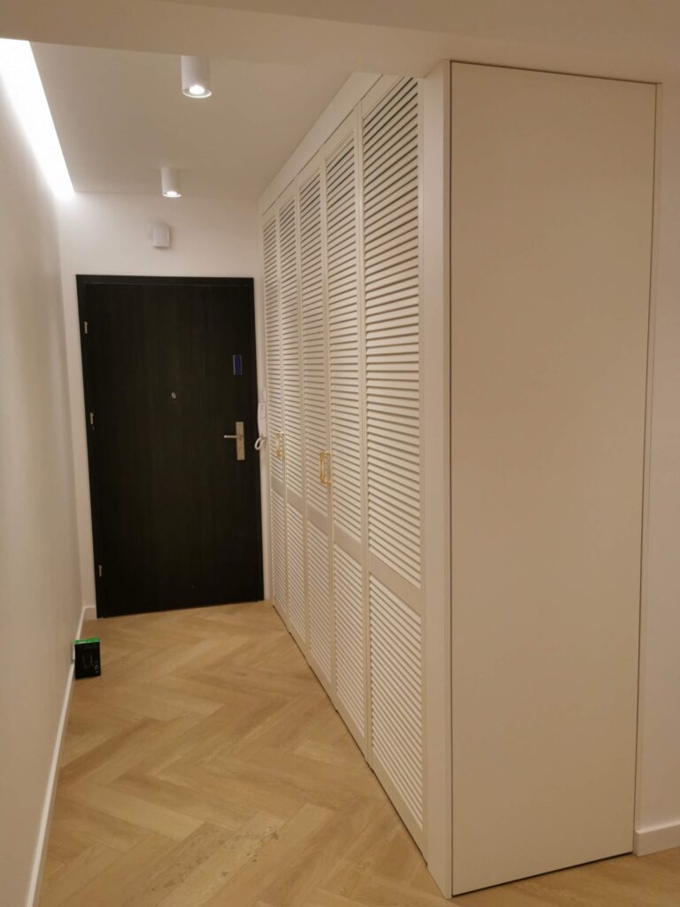 Wysoka biała szafa z lamelowymi drzwiami i złotymi uchwytami