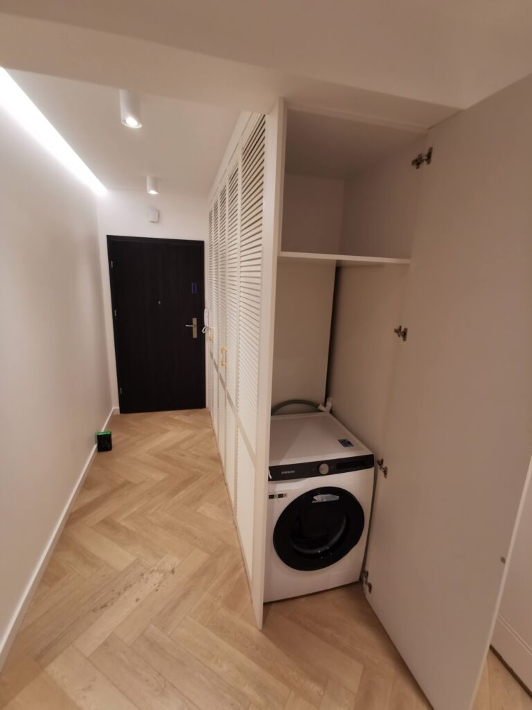 Pralka ukryta za otwartymi drzwiami białej zabudowy w korytarzu